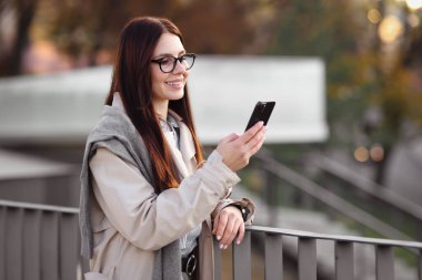 Şık takım elbiseli, açık havada akıllı telefonlu gülümseyen bir iş kadını.