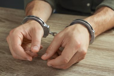 Man in metal handcuffs at wooden table, closeup clipart