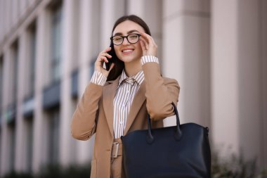 Şık takım elbiseli gülümseyen iş kadını dışarıda akıllı telefondan konuşuyor.