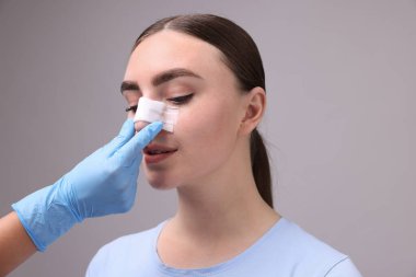 Gri arkaplandaki estetik ameliyattan sonra hastanın burnunu kontrol eden doktor.