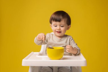 Sarı arka planda, yüksek sandalyede sağlıklı bebek mamaları yiyen şirin bir çocuk.