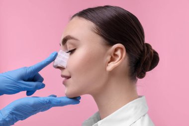 Doktor pembe arka plandaki estetik ameliyattan sonra hastanın burnunu kontrol ediyor.