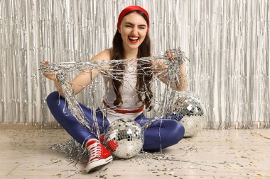 Kapalı alanda gümüş folyo perdelere karşı süslemeli retro kıyafetli duygusal genç kadın.