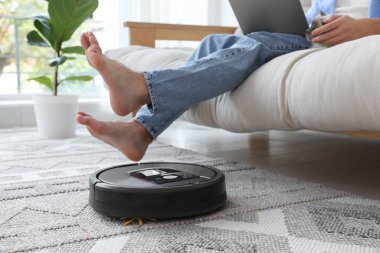Genç adam dizüstü bilgisayar kullanırken elektrikli süpürge evde halı süpürüyor.