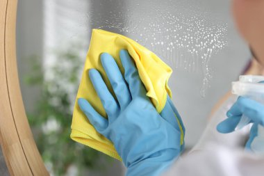 Kadın aynayı kapalı alanda bez parçasıyla silerken temizlik malzemesi kullanıyor.