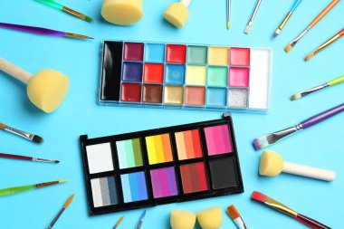 Açık mavi arka planda parlak yüz boyaları, fırçalar ve süngerler, düz yüz.