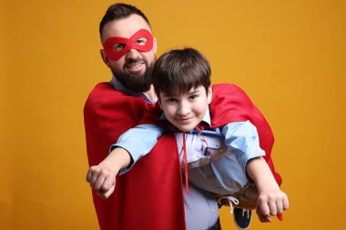 Baba ve oğlu turuncu arka planda süper kahraman kostümleri giyiyorlar.