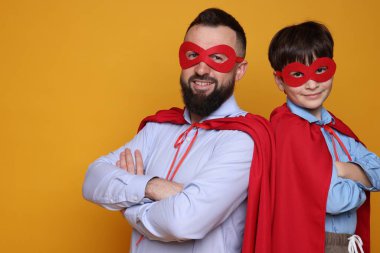 Baba ve oğlu turuncu arka planda süper kahraman kostümleri giyiyorlar, mesaj için yer var.