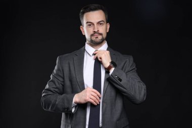Klasik takım elbiseli kendine güvenen adam siyah arka planda kravat düzeltiyor.
