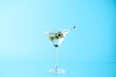 Açık mavi arka planda zeytinli martini kokteyli.