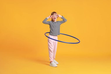 Turuncu arka planda hula hoop egzersizi yaparken V işaretleri gösteren sevimli küçük kız.