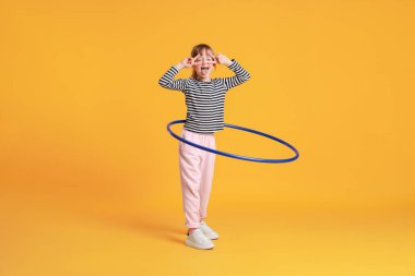 Cute little girl showing V-signs while exercising with hula hoop on orange background clipart