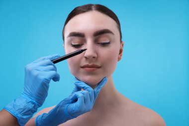 Doktor açık mavi arkaplana karşı estetik ameliyat için hastanın burnuna işaretler çiziyor.