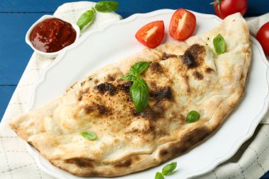 Fesleğenli, domatesli ve mavi ahşap masadaki soslu leziz bir calzone.