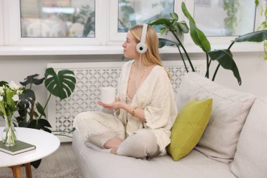 Feng shui. Kulaklığı ve içkisi olan genç bir kadın evdeki kanepede meditasyon yapıyor.