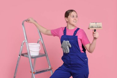 Açık pembe arka planda fırçası, boyası ve merdiveni olan profesyonel bir ressam.