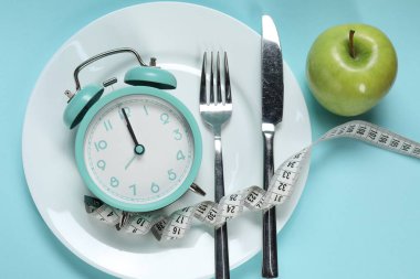 Alarm saati, elma, mezura, açık mavi zemin üzerindeki tabak ve çatal bıçak takımı.