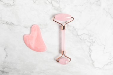 Beyaz mermer zemin üzerinde yüz silindiri ve gua sha aracı, üst görünüm