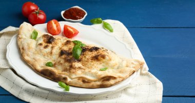 Fesleğenli, domatesli ve mavi ahşap masadaki soslu leziz calzone, yakın plan.