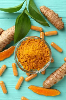 Turkuaz ahşap masa üzerinde zerdeçal kapsülleri, kökler, toz ve yeşil yapraklar.