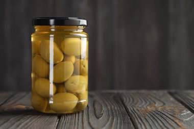 Cam kavanozda yeşil zeytin turşusu, ahşap masada gri arka planda, metin için boşluk.