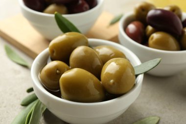 Lezzetli marine edilmiş zeytinler kaselerde ve yeşil yapraklar gri masada, yakın plan.