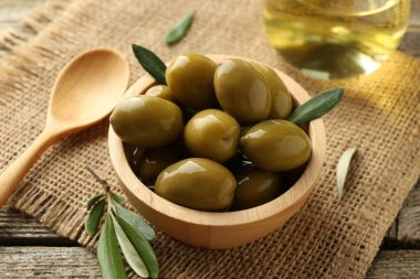 Lezzetli zeytinler kasede, yeşil yapraklar, kaşık ve yağ ahşap masada, yakın plan.