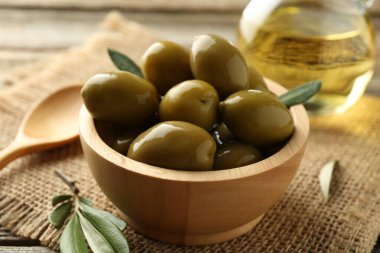 Lezzetli zeytinler kasede, yeşil yapraklar, masada kaşık ve yağ, yakın plan.