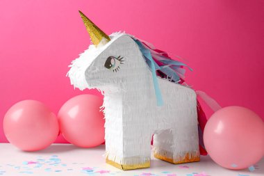 Tek boynuzlu at şeklinde parlak pinyata ve pembe arka planda beyaz masada balonlar.
