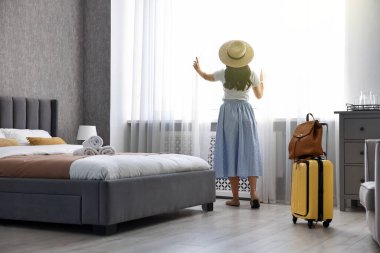 Otel odasındaki perdeleri açan bavullu gezgin.
