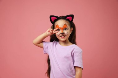 Pembe arka planda barış işareti gösteren yüzü ve kulağı boyalı sevimli bir kız.