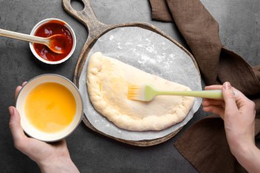 Gri masadaki calzone pizzasında yumurta sarısı fırçalayan kadın.