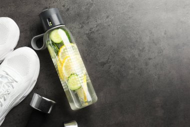 Şişedeki limonlu su, gri yüzeyde spor ayakkabılar ve dambıllar, düz uzanmış. Metin için boşluk