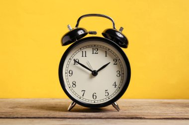 Sarı arka planda, ahşap masada siyah bir çalar saat.