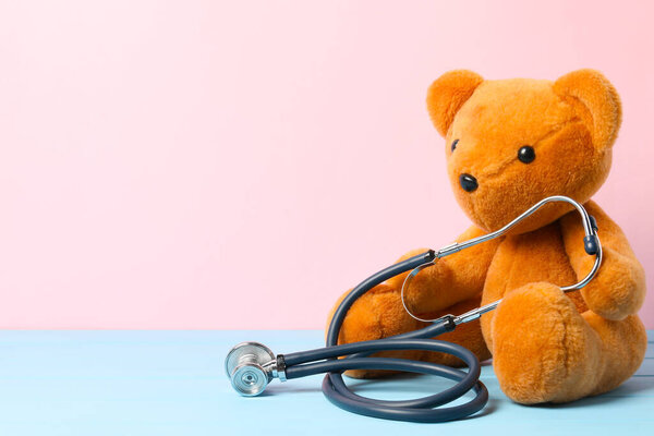 Pediatrics concept. Teddy bear with stethoscope on light blue wooden table against pink background, space for text