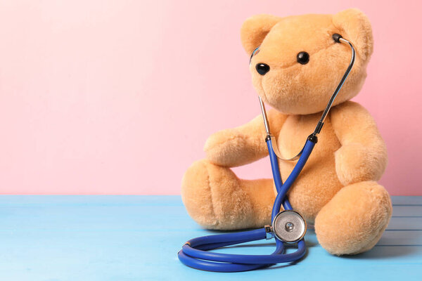 Pediatrics concept. Teddy bear with stethoscope on light blue wooden table against pink background, space for text