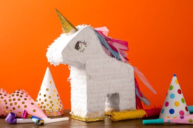 Tek boynuzlu at şeklindeki parlak pinyata ve turuncu arka plandaki parti aksesuarları.