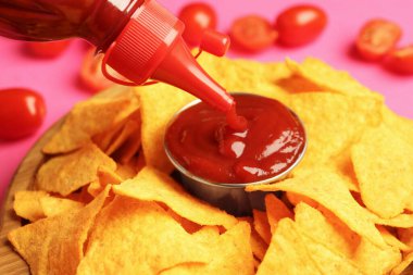 Pouring tasty ketchup in bowl with nachos on pink background, closeup clipart