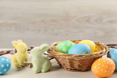 Hasır sepette birçok süslü Paskalya yumurtası, söğüt dalları ve ahşap masada dekoratif tavşanlar, yakın plan.