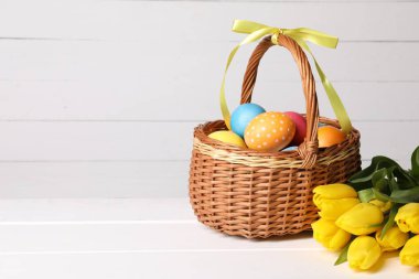Hasır sepetteki Paskalya yumurtaları ve beyaz ahşap masadaki güzel laleler, metin için yer.