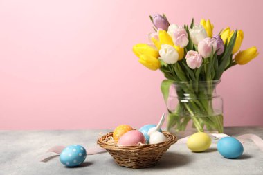 Hasır sepetteki Paskalya yumurtaları, tavşan figürü ve pembe arka planda gri masadaki güzel laleler.