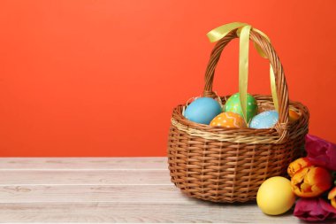 Hasır sepetteki Paskalya yumurtaları ve ahşap masadaki güzel laleler turuncu arka planda, metin için yer var.