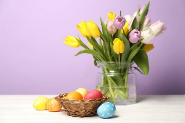 Hasır sepette paskalya yumurtaları ve beyaz ahşap masada leylak arka planda güzel laleler.
