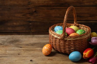 Hasır sepetteki Paskalya yumurtaları ve ahşap masada güzel laleler, metin için yer.