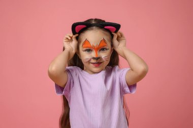 Pembe arka planda kedi gibi yüzü kulakları boyalı tatlı bir kız.