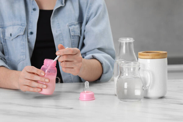 Mother making baby formula in feeding bottle at table indoors, closeup