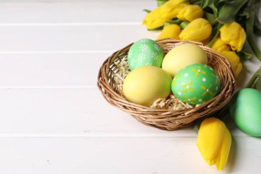 Hasır sepetteki Paskalya yumurtaları ve beyaz ahşap masadaki güzel laleler, yakın plan. Metin için boşluk