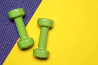 Sarı arka planda dambıllar ve fitness paspası, düz yatış. Metin için boşluk