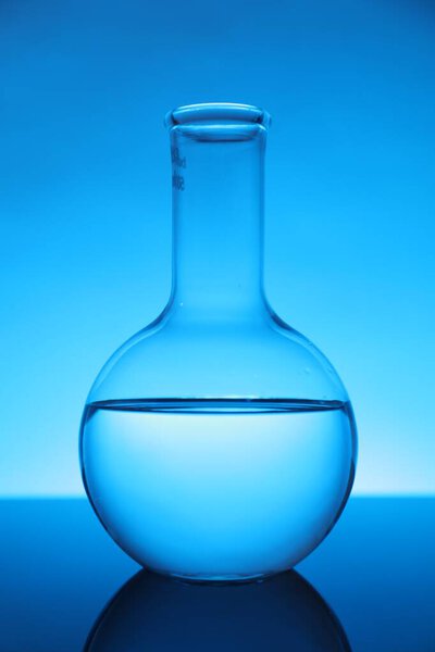 Flask with liquid on mirror surface against light blue background. Laboratory equipment
