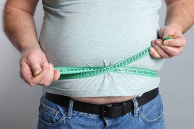 Kilolu bir adam, göbeğini gri arka planda bantla ölçüyor.